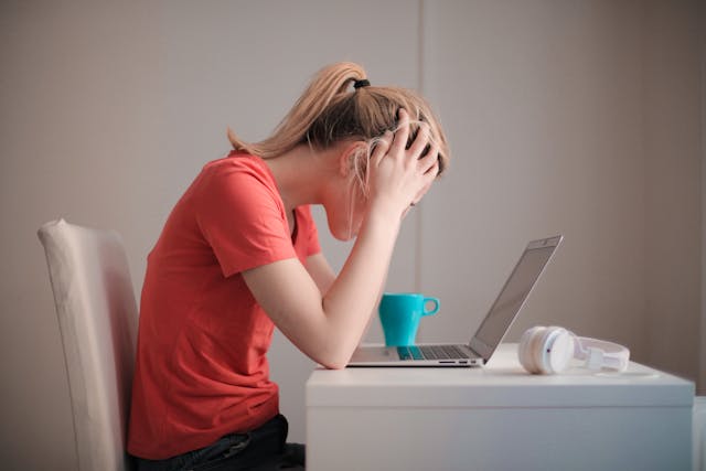 A person with long hair sitting at a table in front of a laptop, with their head in their hands.